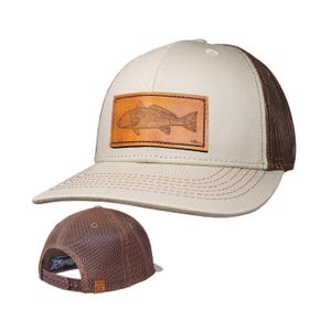 Op de afbeelding: Een beige en bruine truckerpet met een rechthoekige leren patch op de voorkant met een gedetailleerde gegraveerde illustratie van een vis. De pet heeft een beige voorpaneel, een bruine mesh achterkant en een verstelbare plastic snapbacksluiting.