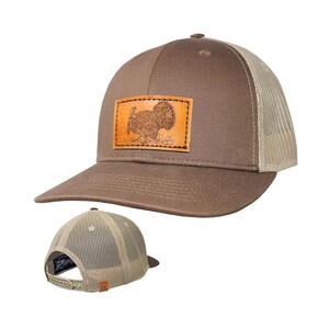May include: A brown baseball cap with a tan mesh back and an adjustable snapback closure. The front features a rectangular tan leather patch with a detailed engraved illustration of a turkey. The cap has a structured crown and a curved brim.