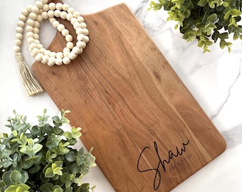 May include: A wooden cutting board with a natural wood grain finish. The board has a handle and is engraved with the text "Shaw Jim & Kelly".
