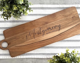 May include: A long, rectangular wooden serving board with a handle. The board is engraved with the text "Montgomery Blair + Polly" in a cursive font.