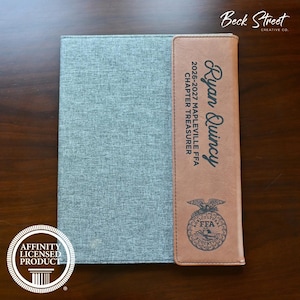 May include: A gray fabric-covered portfolio featuring a vertical tan faux leather accent strip on the right side. The strip is engraved with the name Ryan Quincy, the text 2026-2027 MAPLEVILLE FFA CHAPTER TREASURER, and an FFA emblem logo.