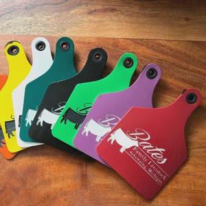 May include: A collection of colorful livestock ear tags in various shades, including yellow, white, teal, black, green, purple, and red. Each tag features a white silhouette of a cow and the name "Bates Family Livestock" in white text.