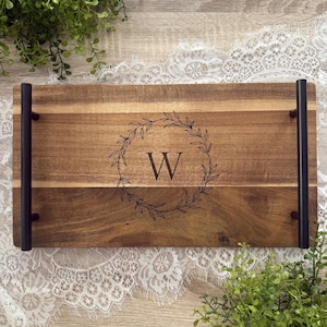 May include: A wooden serving tray with black metal handles. The tray has a natural wood finish and features a laser-engraved wreath with the letter "W" in the center.