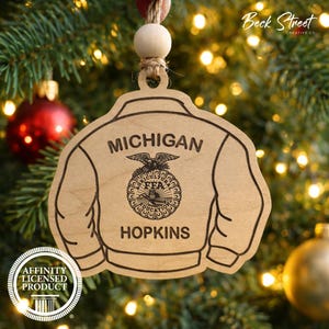 May include: A wooden ornament shaped like a jacket, with the words "MICHIGAN" and "HOPKINS" engraved on it. The ornament features the FFA emblem and is hanging from a Christmas tree. The ornament is a licensed product.