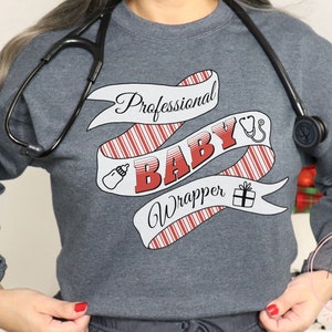 May include: A gray sweatshirt with a red and white candy cane stripe design. The text "Professional Baby Wrapper" is written on the design, along with a stethoscope, a baby bottle, and a gift box.