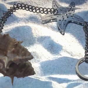 May include: A silver tea infuser shaped like a starfish with a chain and a ring attached. The infuser is on a white sandy surface with a brown seashell.