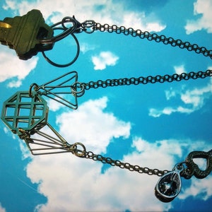 Puede incluir: Un llavero plateado con un dije geométrico, un dije de llave en forma de corazón y un dije de llave tradicional. La cadena cuelga contra un cielo azul con nubes blancas.