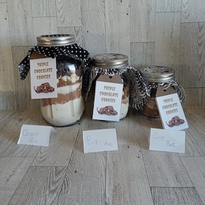 Op de afbeelding: Drie glazen potten gevuld met gelaagde ingrediënten voor triple chocolate cookies. Elke pot heeft een deksel en stof, en een label met de tekst "Triple Chocolate Cookies". De potten zijn gelabeld Quart (946ml), Pint (473ml) en Cup (237ml).