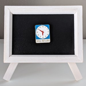 May include: A white framed chalkboard with a blue and white enamel pin featuring a clock face with red hands and the text "WILL RETURN".