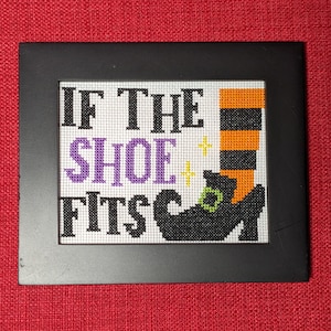 May include: A black framed cross-stitch with the text "If the shoe fits" and a witch's shoe with green and orange accents.