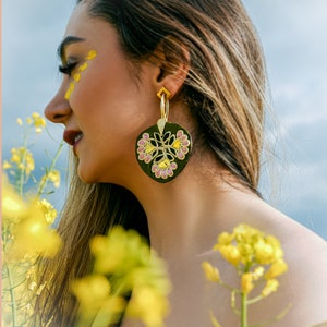 May include: A woman with long brown hair wearing a pair of large, gold hoop earrings with a black and pink floral design. The earrings are hanging from her earlobes and are visible against a backdrop of yellow flowers.