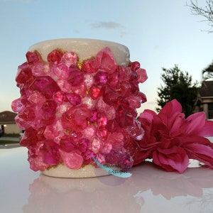 May include: White ceramic mug covered in pink and clear glitter-like gems. The mug has a gold glitter band around the top edge. The text "Handmade By CATTLYNN" is written in blue on the bottom right side of the mug.