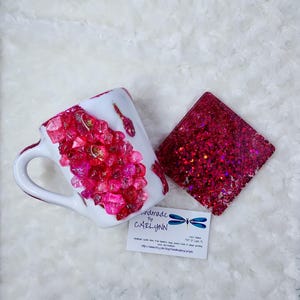 May include: A white ceramic mug and a square coaster, both adorned with vibrant pink and red crystal-like embellishments and gold accents. The mug features a handle and the coaster is covered in glitter. A business card is also present.