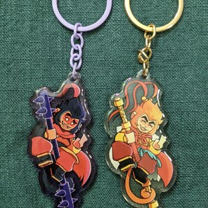 以下が含まれることがあります： 漫画の猿のキャラクターが描かれたアクリルキーホルダー2点。左のキーホルダーはラベンダー色のリングで、トゲ付きの棍棒を持った黒髪の猿が描かれています。右のキーホルダーはゴールドのリングで、杖を持った金髪の猿が描かれています。