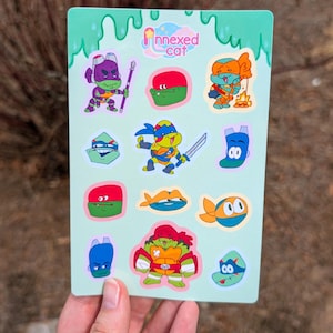 May include: A light green sticker sheet featuring various cartoon illustrations of turtle characters in masks, including full-body poses and headshots. The top center displays the text "Annexed cat" with a small star icon. The designs include vibrant colors like purple, red, orange, and blue.