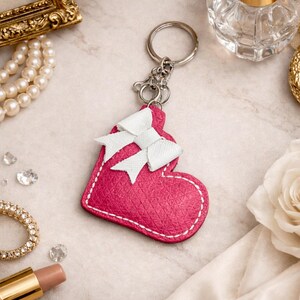 May include: A pink heart-shaped keychain with a white bow detail. The keychain has a silver ring and clasp. The heart has white stitching around the edges. The background includes jewelry and a perfume bottle.