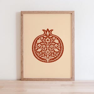 May include: Framed art print featuring a detailed pomegranate design in shades of red and cream. The botanical illustration is centered on a cream-colored background, encased in a wooden frame. The artwork is displayed on a wooden surface.