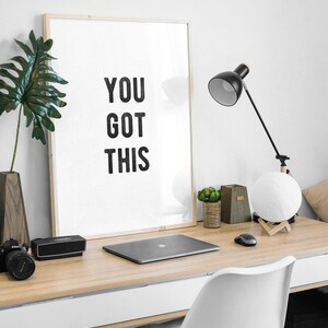 May include: A white framed poster with the text "YOU GOT THIS" on a wooden desk with a laptop, a lamp, and a potted plant.