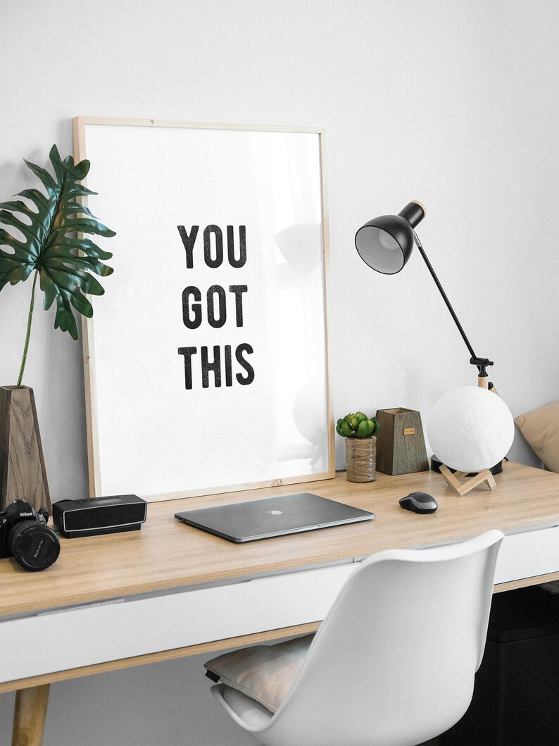 May include: A white framed poster with the text "YOU GOT THIS" on a wooden desk with a laptop, a lamp, and a potted plant.