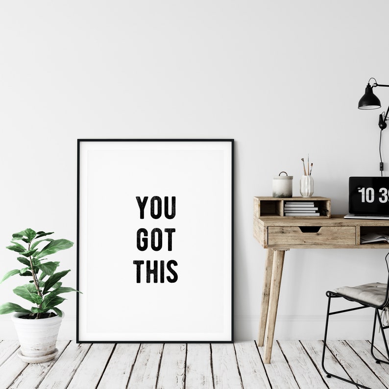 May include: A black framed poster with the text "YOU GOT THIS" in white letters on a white background. The poster is hanging on a white wall above a wooden desk with a laptop, a clock, and a plant.
