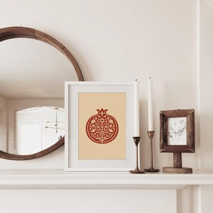 May include: A framed print featuring a red pomegranate design with floral details on a cream background. The artwork is displayed on a white mantel, alongside candles and a small framed picture. A large, round mirror is visible in the background.