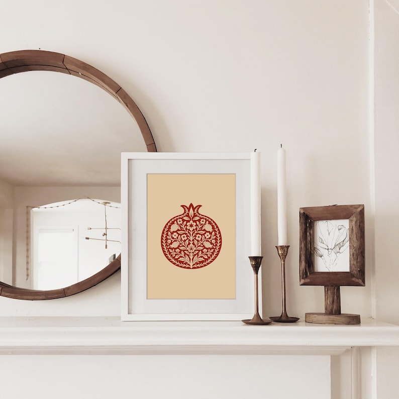 May include: A framed print featuring a red pomegranate design with floral details on a cream background. The artwork is displayed on a white mantel, alongside candles and a small framed picture. A large, round mirror is visible in the background.