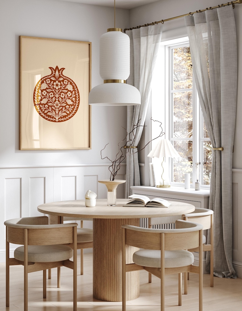 May include: A dining room scene featuring a round wooden table with four chairs. A framed artwork with a pomegranate design hangs on the wall. A white pendant light hangs above the table, and a window with sheer curtains is visible.