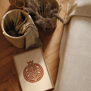 May include: A framed print featuring a stylized pomegranate design in shades of brown and cream. Woven baskets and a folded blanket are visible, along with dried plants. The scene is set on a wooden floor, suggesting a home decor theme.