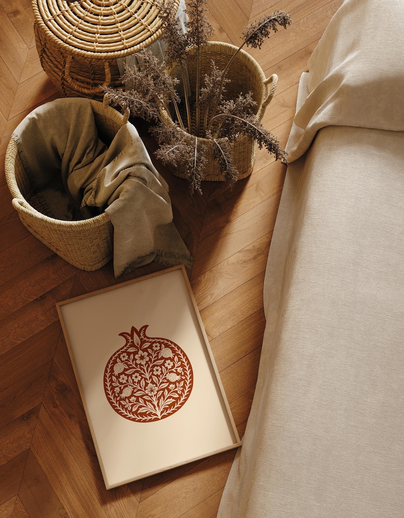 May include: A framed print featuring a stylized pomegranate design in shades of brown and cream. Woven baskets and a folded blanket are visible, along with dried plants. The scene is set on a wooden floor, suggesting a home decor theme.