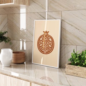 May include: A framed art print featuring a stylized pomegranate design in shades of brown and cream. The artwork is displayed on a countertop with other decorative items, including a white pitcher, wooden bowls, and a small plant in a wooden planter.