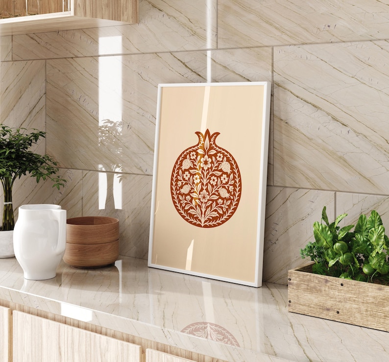 May include: A framed art print featuring a stylized pomegranate design in shades of brown and cream. The artwork is displayed on a countertop with other decorative items, including a white pitcher, wooden bowls, and a small plant in a wooden planter.