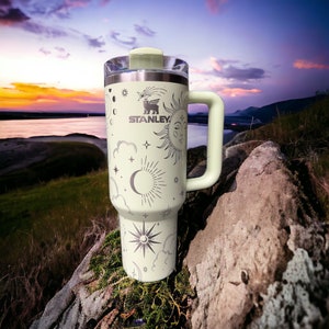 K&ouml;nnte beinhalten: Eine hellgr&uuml;ne Edelstahl-Reisebecher mit schwarzem Griff und einem wei&szlig;en zellischen Design. Der Becher zeigt einen Halbmond, Sterne und eine Sonne mit einem Gesicht. Der Text "STANLEY" ist auf dem Becher aufgedruckt.