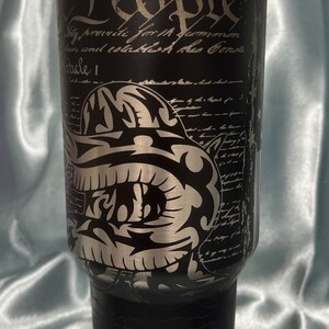 Puede incluir: Un vaso de acero inoxidable negro con un gr&aacute;fico plateado de una calavera con casco y el texto "We the People" y "Tread" en la parte inferior. El vaso tiene una tapa negra y una pajita plateada.