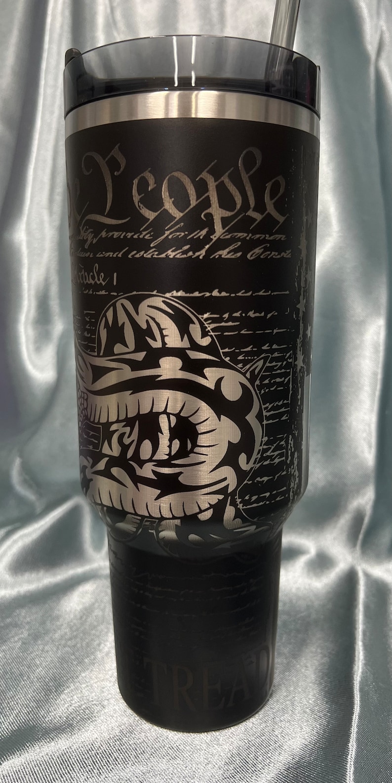 Puede incluir: Un vaso de acero inoxidable negro con un gr&aacute;fico plateado de una calavera con casco y el texto "We the People" y "Tread" en la parte inferior. El vaso tiene una tapa negra y una pajita plateada.