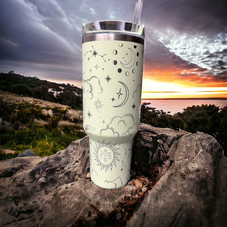K&ouml;nnte beinhalten: Ein Edelstahlbecher mit hellgr&uuml;nem Hintergrund und einem schwarz-wei&szlig;en Design von Himmelsk&ouml;rpern, darunter Sterne, Monde und eine Sonne mit einem Gesicht. Der Becher steht auf einem Stein mit einem Sonnenuntergang im Hintergrund.