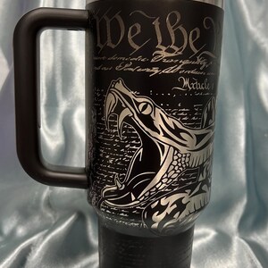 Puede incluir: Un vaso de acero inoxidable negro con una manija negra y una tapa negra. El vaso tiene una serpiente plateada con colmillos y el texto "We the People" y "Article 1" en el lateral. La parte inferior del vaso dice "Don't Tread on Me".