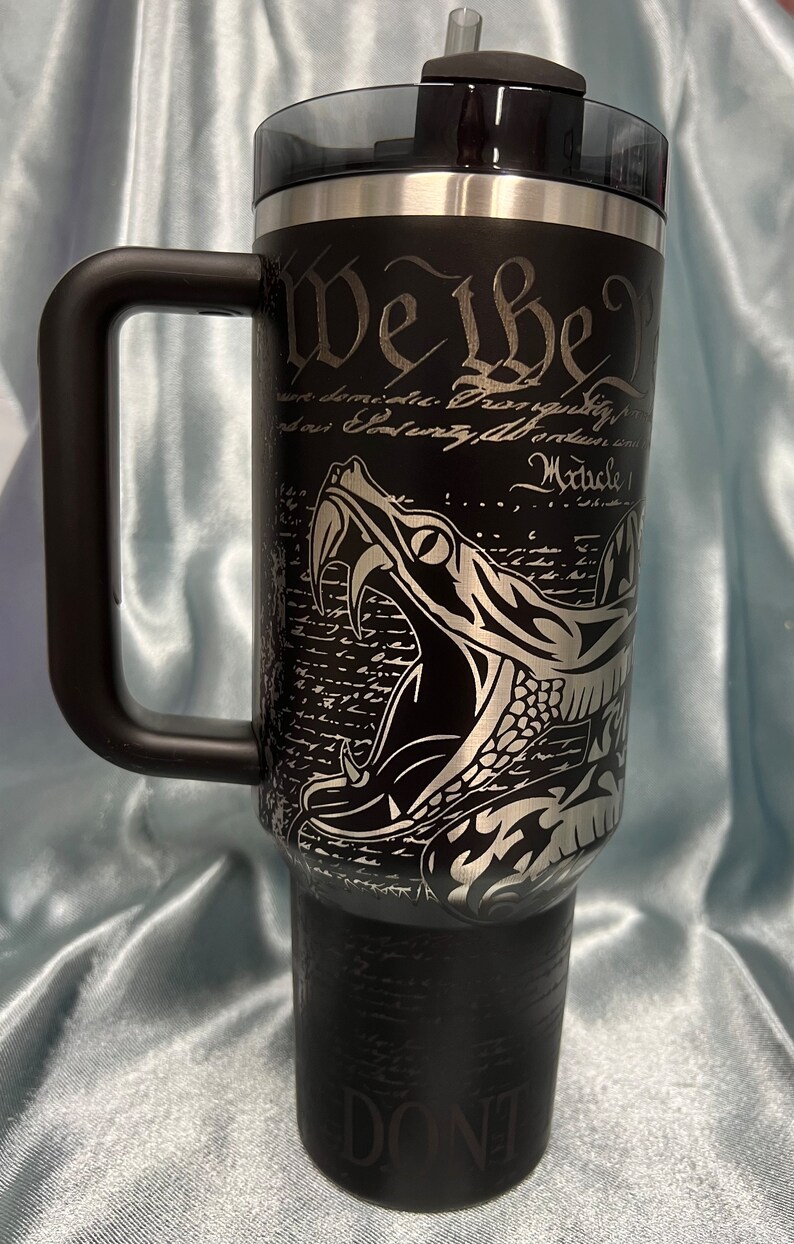 Puede incluir: Un vaso de acero inoxidable negro con una manija negra y una tapa negra. El vaso tiene una serpiente plateada con colmillos y el texto "We the People" y "Article 1" en el lateral. La parte inferior del vaso dice "Don't Tread on Me".