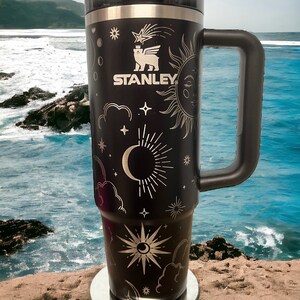 K&ouml;nnte beinhalten: Schwarze Edelstahl-Reisebecher mit goldenem Himmelsdesign, einschlie&szlig;lich einer Mondsichel, Sternen und einer Sonne. Der Becher hat einen schwarzen Griff und das Stanley-Logo ist auf der Seite aufgedruckt.