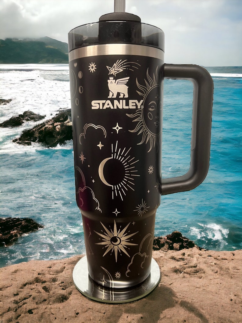 K&ouml;nnte beinhalten: Schwarze Edelstahl-Reisebecher mit goldenem Himmelsdesign, einschlie&szlig;lich einer Mondsichel, Sternen und einer Sonne. Der Becher hat einen schwarzen Griff und das Stanley-Logo ist auf der Seite aufgedruckt.