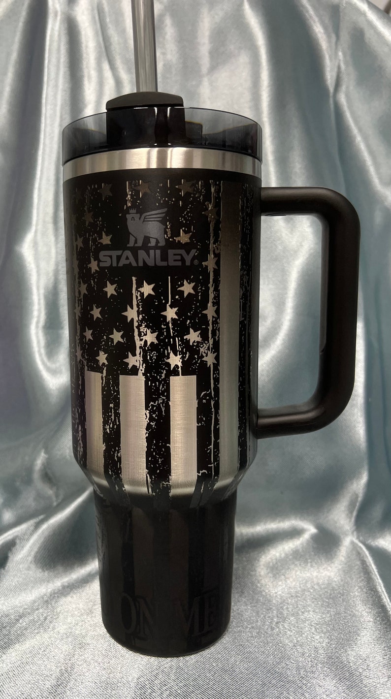 Puede incluir: Una taza de viaje de acero inoxidable negra y plateada con un asa negra y un dise&ntilde;o de bandera estadounidense desgastado. La taza tiene el logotipo de Stanley y las palabras "On Me" impresas en la parte inferior.