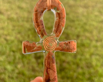 May include: Ankh pendant with a copper wire wrap. The ankh is filled with small, reddish-brown stones and clear resin. The pendant is held in a hand against a blurred green background.