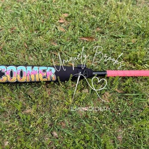 Puede incluir: Un bate de b&eacute;isbol negro con un agarre rosa y un dise&ntilde;o personalizado "COOMER" en colores arco&iacute;ris en el ca&ntilde;&oacute;n.