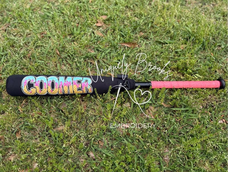 Puede incluir: Un bate de b&eacute;isbol negro con un agarre rosa y un dise&ntilde;o personalizado "COOMER" en colores arco&iacute;ris en el ca&ntilde;&oacute;n.