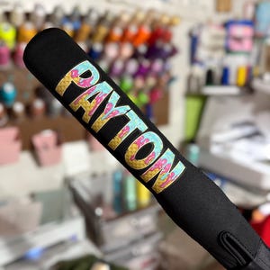 Puede incluir: Empu&ntilde;adura de bate de b&eacute;isbol negro con un dise&ntilde;o de helado colorido que dice "PAYTON" en letras doradas.