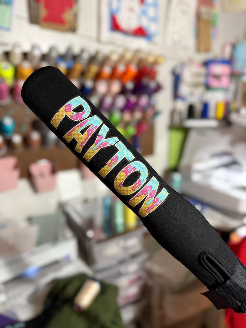 Puede incluir: Empu&ntilde;adura de bate de b&eacute;isbol negro con un dise&ntilde;o de helado colorido que dice "PAYTON" en letras doradas.