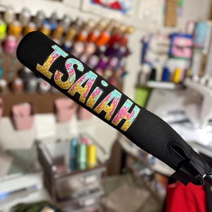 Puede incluir: Una funda de bate de b&eacute;isbol negra con un dise&ntilde;o de cono de helado colorido y el nombre "ISAIAH" escrito en letras amarillas.