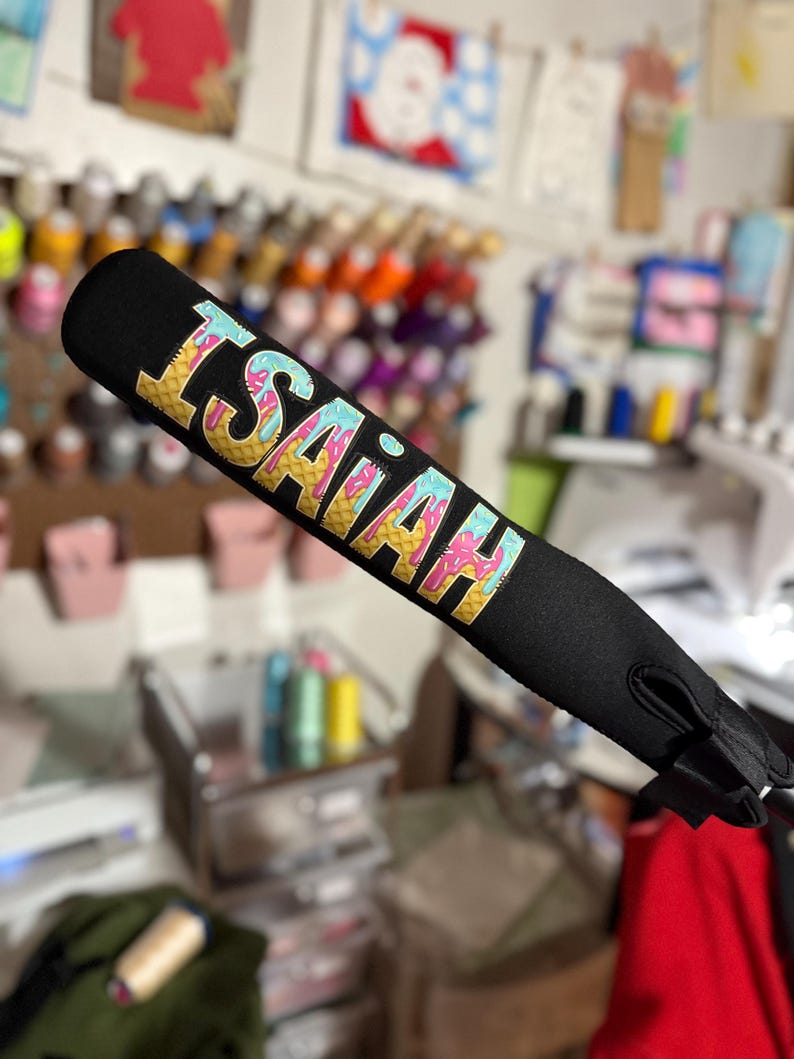 Puede incluir: Una funda de bate de b&eacute;isbol negra con un dise&ntilde;o de cono de helado colorido y el nombre "ISAIAH" escrito en letras amarillas.
