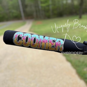 Puede incluir: Un bate de b&eacute;isbol negro con un dise&ntilde;o bordado personalizado que dice "COOMER" en una fuente de cono de helado que gotea con colores rosa, azul y amarillo. El bate tambi&eacute;n est&aacute; etiquetado como "Uniquely Brooks Embroidery".