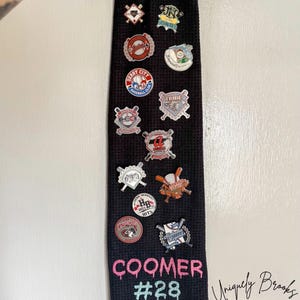 May include: Black golf towel with a white embroidered name "COOMER #28" and various baseball team pins attached. The towel has a waffle texture and a loop for hanging. The text "Uniquely Brooks Embroidery" is embroidered in white on the bottom right corner.