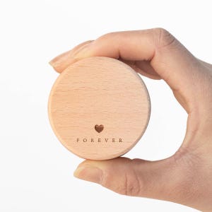 以下が含まれることがあります： 小さなハートと「FOREVER」の文字が刻まれた、丸い明るい色の木製オブジェクト。白い背景を背景に、手に持たれています。蓋または小さな容器のように見えます。